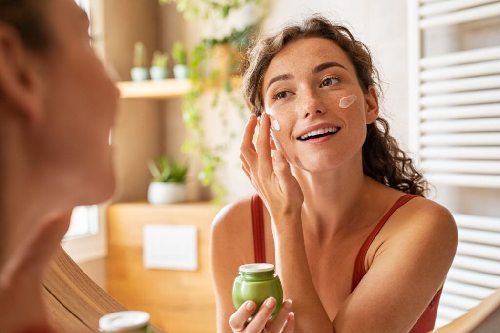 A woman with curly hair and freckles applies a cream to her face while looking at herself in a mirror. She holds a green jar of skincare product in one hand and smiles, enjoying her skincare routine. The background features a cozy bathroom setting with natural wooden elements and potted plants, creating a fresh and calming atmosphere.