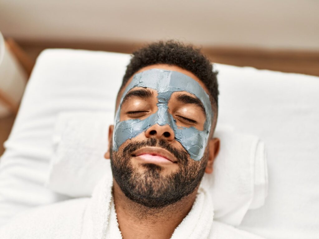 A man with a neatly trimmed beard is lying down with his eyes closed, enjoying a relaxing skincare treatment. He is wearing a gray facial mask that covers his forehead, nose, and cheeks. Dressed in a white robe and resting on a towel, he appears calm and rejuvenated, in a serene spa-like setting.